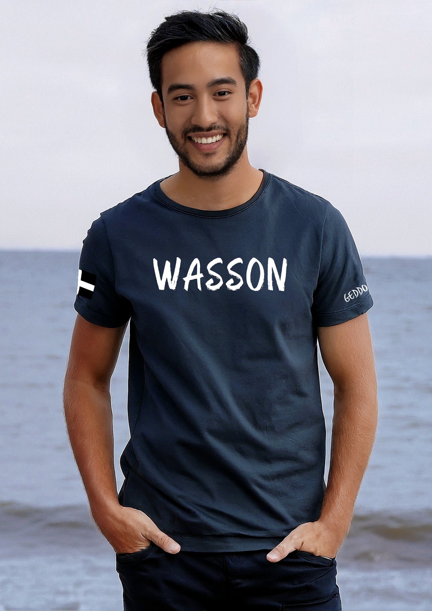 Model wearing a navy t-shirt with "WASSON" in bold white text on the chest, featuring a Cornish flag on the sleeve.